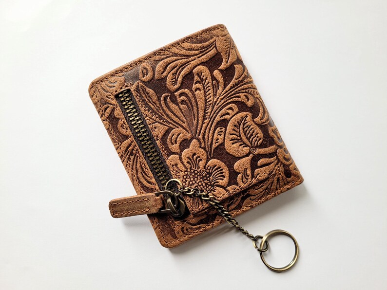 May include: Brown leather wallet with a floral embossed design. The wallet has a zipper closure and a key ring attached with a chain. The wallet is rectangular and appears to be new.
