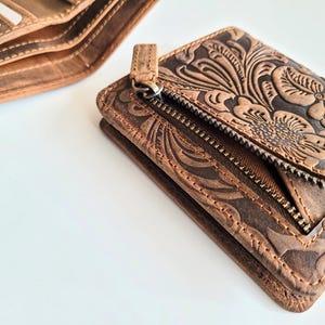 May include: Close-up of two brown leather wallets. One wallet is open, revealing card slots. The other wallet is a small, zippered cardholder with a floral embossed design. The leather has a distressed finish.