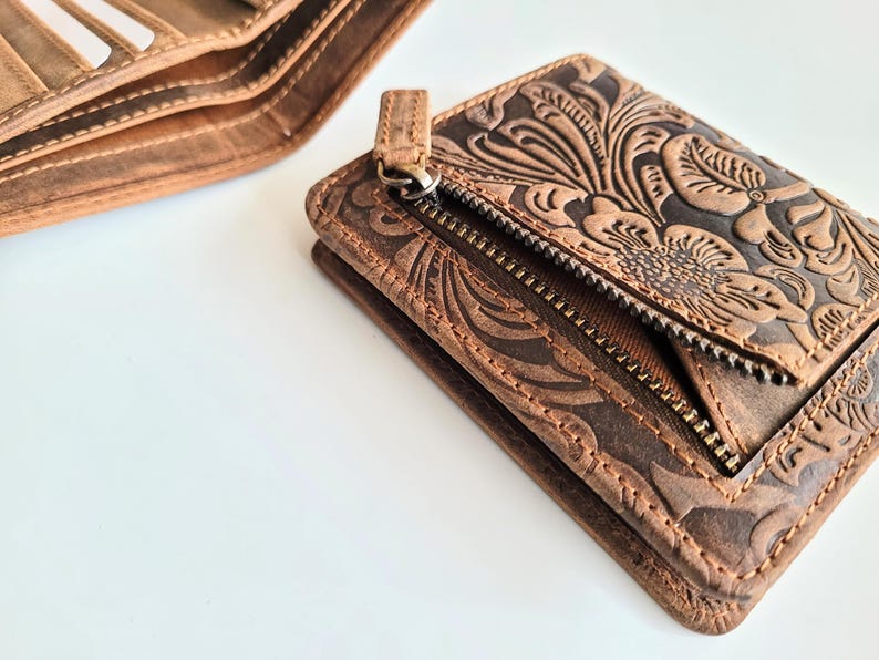 May include: Close-up of two brown leather wallets. One wallet is open, revealing card slots. The other wallet is a small, zippered cardholder with a floral embossed design. The leather has a distressed finish.