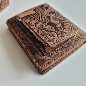 May include: A brown leather wallet with a floral embossed design. The wallet has a zippered compartment and is stitched around the edges. The leather has a distressed look, giving it a vintage feel. The wallet is a bi-fold style.