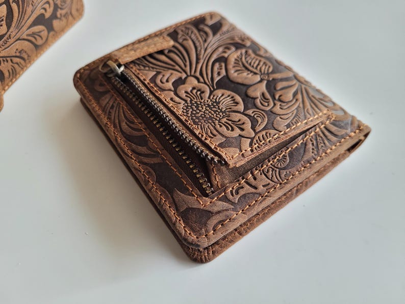 May include: A brown leather wallet with a floral embossed design. The wallet has a zippered compartment and is stitched around the edges. The leather has a distressed look, giving it a vintage feel. The wallet is a bi-fold style.