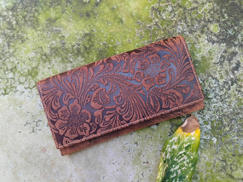 K&ouml;nnte beinhalten: Eine braune Ledergeldb&ouml;rse mit einem gepr&auml;gten Blumenmuster. Die Geldb&ouml;rse hat einen Klappverschluss und ist aus weichem, geschmeidigem Leder gefertigt.