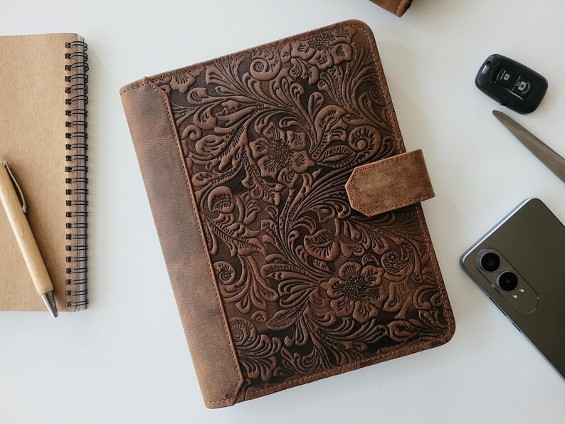 Puede incluir: Un diario de cuero marr&oacute;n con un dise&ntilde;o floral en relieve y un cierre de correa. Un cuaderno de espiral y un bol&iacute;grafo son visibles a la izquierda. Una llave de coche, un cuchillo y un tel&eacute;fono inteligente tambi&eacute;n est&aacute;n en la imagen.