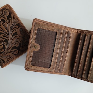 May include: Two brown leather wallets. One is closed, featuring a floral embossed design. The other is open, revealing card slots and a clear ID window. Both wallets have visible stitching and a worn, rustic appearance.