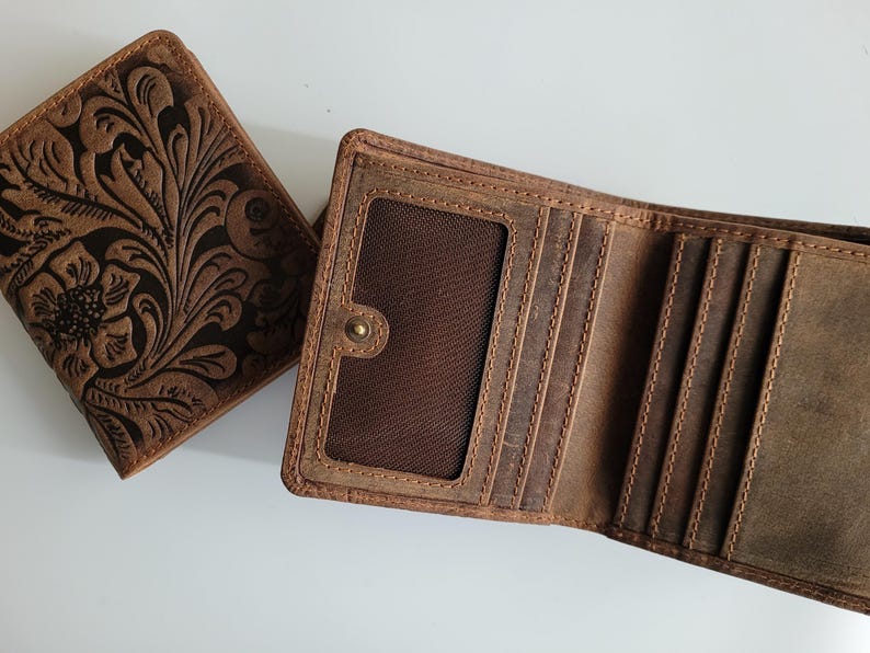 May include: Two brown leather wallets. One is closed, featuring a floral embossed design. The other is open, revealing card slots and a clear ID window. Both wallets have visible stitching and a worn, rustic appearance.