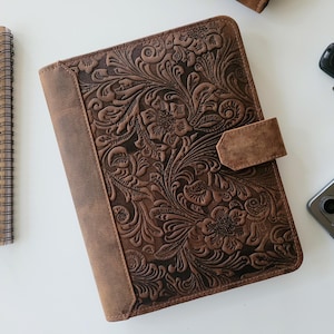 May include: A brown leather planner with a floral embossed design. The planner has a flap closure and a brown leather strap.