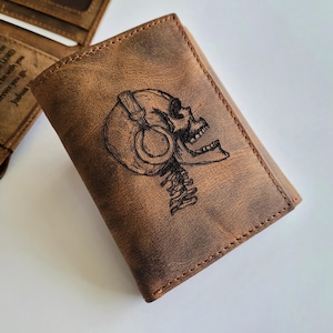 May include: A brown leather trifold wallet with a skull wearing headphones engraved on the front. The wallet has a distressed look and is stitched with brown thread.