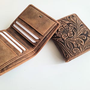May include: Two brown leather wallets. One is open, revealing card slots and a bill compartment. The other wallet is closed, featuring an embossed floral design. Both wallets have a snap closure and a distressed finish, suggesting a vintage or rustic style.