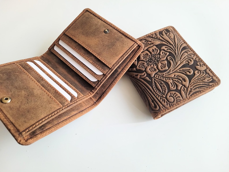 May include: Two brown leather wallets. One is open, revealing card slots and a bill compartment. The other wallet is closed, featuring an embossed floral design. Both wallets have a snap closure and a distressed finish, suggesting a vintage or rustic style.