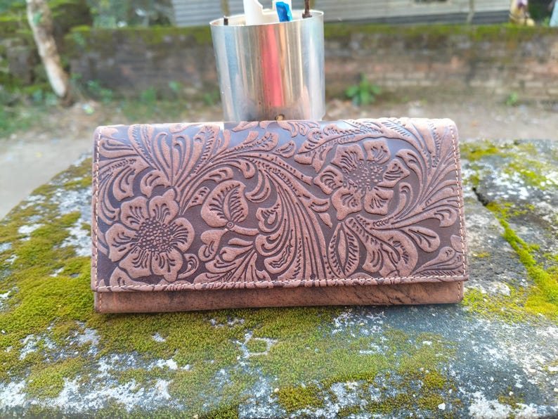 K&ouml;nnte beinhalten: Eine braune Ledergeldb&ouml;rse mit einem gepr&auml;gten Blumenmuster. Die Geldb&ouml;rse hat einen Klappverschluss und ist aus weichem, geschmeidigem Leder gefertigt.
