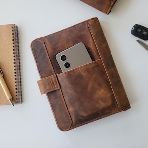 Puede incluir: Una funda de tableta de cuero marr&oacute;n con un tel&eacute;fono en el bolsillo delantero. Un cuaderno de espiral, un bol&iacute;grafo, un llavero de coche y unas tijeras tambi&eacute;n est&aacute;n sobre la superficie blanca. El cuero tiene un aspecto envejecido.