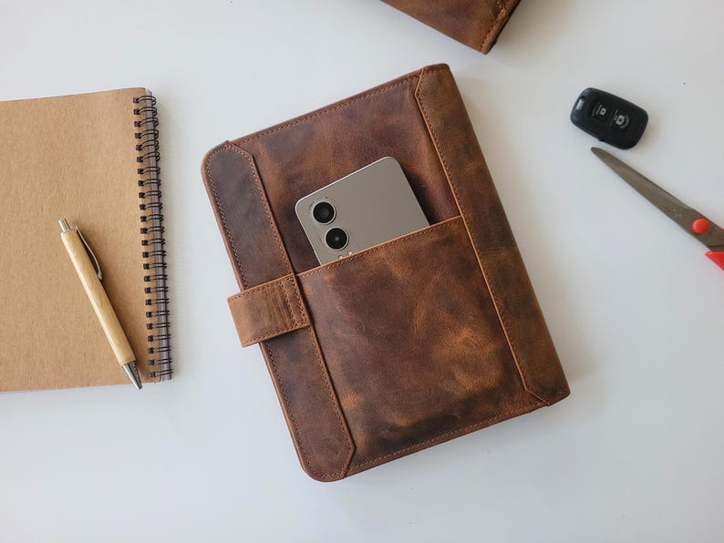 Puede incluir: Una funda de tableta de cuero marr&oacute;n con un tel&eacute;fono en el bolsillo delantero. Un cuaderno de espiral, un bol&iacute;grafo, un llavero de coche y unas tijeras tambi&eacute;n est&aacute;n sobre la superficie blanca. El cuero tiene un aspecto envejecido.