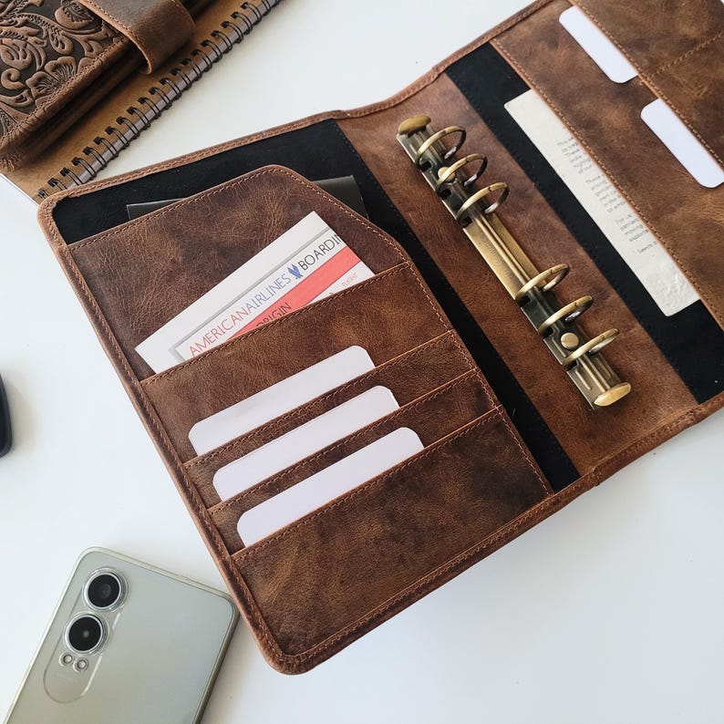 Puede incluir: Carpeta de cuero marr&oacute;n abierta con m&uacute;ltiples ranuras para tarjetas y un mecanismo de encuadernaci&oacute;n de anillas met&aacute;licas. La carpeta contiene tarjetas blancas y una tarjeta de embarque de American Airlines. Tambi&eacute;n se ve un smartphone y un cuaderno de cuero.