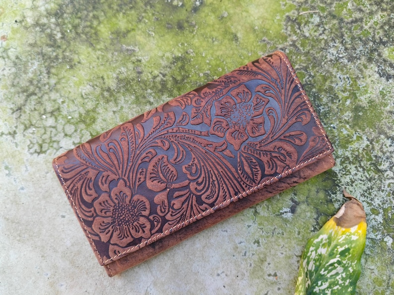 K&ouml;nnte beinhalten: Eine braune Lederb&ouml;rse mit einem gepr&auml;gten Blumenmuster. Die B&ouml;rse hat einen Klappverschluss und ist mit filigranen Blumenmustern verziert.