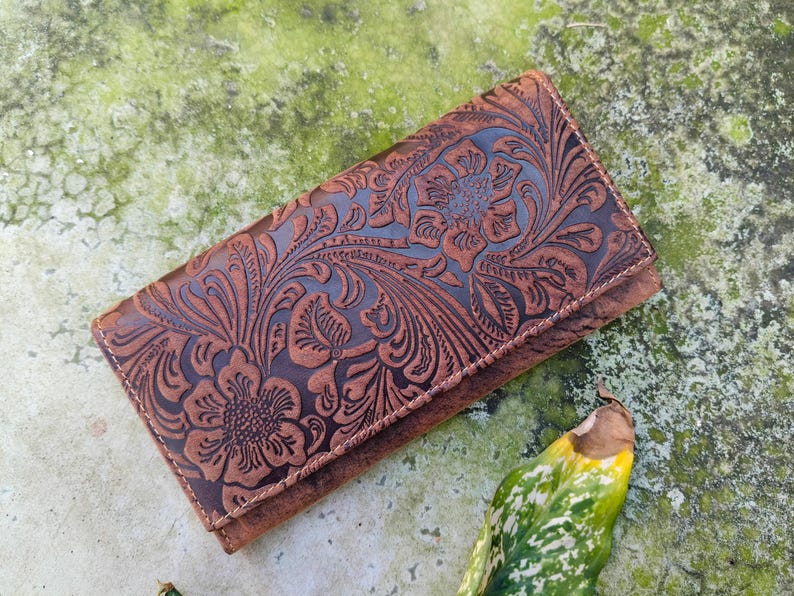 K&ouml;nnte beinhalten: Eine braune Lederb&ouml;rse mit einem gepr&auml;gten Blumenmuster. Die B&ouml;rse hat einen Klappverschluss und ist aus weichem, geschmeidigem Leder gefertigt.