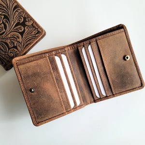 May include: Two brown leather wallets. One is open, revealing card slots and a snap closure. The other wallet is closed, with a floral embossed design. The wallets are made of distressed leather.
