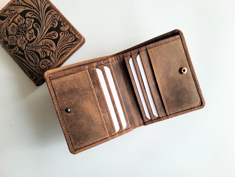 May include: Two brown leather wallets. One is open, revealing card slots and a snap closure. The other wallet is closed, with a floral embossed design. The wallets are made of distressed leather.