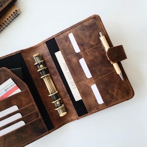 Puede incluir: Agenda de cuero marr&oacute;n abierto con m&uacute;ltiples bolsillos y un portal&aacute;pices. El planificador tiene un mecanismo de encuadernaci&oacute;n de color lat&oacute;n y un bol&iacute;grafo de madera. Varias tarjetas de &iacute;ndice blancas y una tarjeta de embarque son visibles.