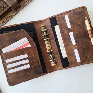 Puede incluir: Agenda de cuero marr&oacute;n abierto con m&uacute;ltiples bolsillos, un bol&iacute;grafo y un mecanismo de encuadernaci&oacute;n. El planificador contiene una tarjeta con el texto "AMERICAN AIRLINES BOARDING". Un cuaderno de cuero a juego es visible en el fondo.