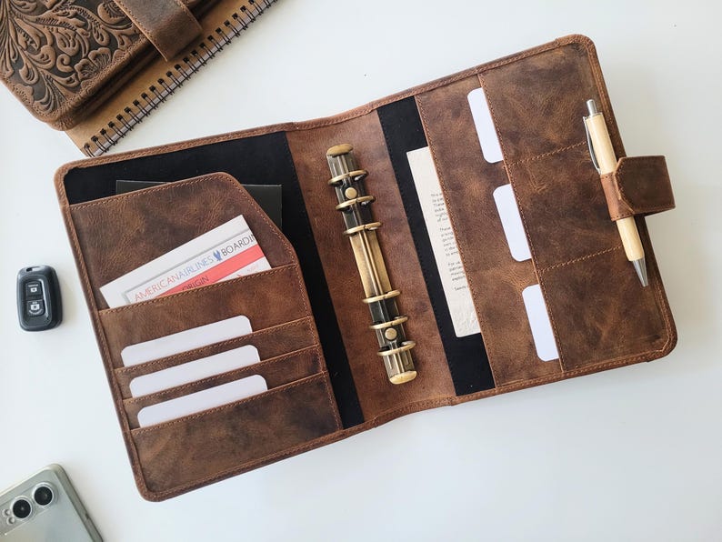 Puede incluir: Agenda de cuero marr&oacute;n abierto con m&uacute;ltiples bolsillos, un bol&iacute;grafo y un mecanismo de encuadernaci&oacute;n. El planificador contiene una tarjeta con el texto "AMERICAN AIRLINES BOARDING". Un cuaderno de cuero a juego es visible en el fondo.