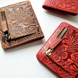May include: Three leather wallets with floral embossed designs. One is brown with a dark zip, and two are red with gold zips. The wallets are rectangular and have a zip pocket on the front.