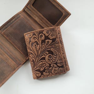 K&ouml;nnte beinhalten: Eine braune Lederb&ouml;rse mit drei Falten und einem gepr&auml;gten Blumenmuster. Die Brieftasche hat mehrere Kartenf&auml;cher und ein Geldfach.