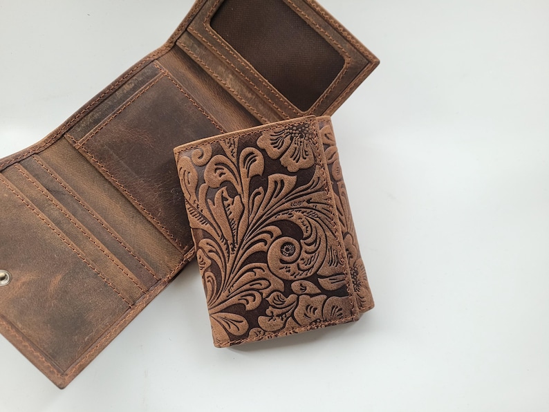K&ouml;nnte beinhalten: Eine braune Lederb&ouml;rse mit drei Falten und einem gepr&auml;gten Blumenmuster. Die Brieftasche hat mehrere Kartenf&auml;cher und ein Geldfach.