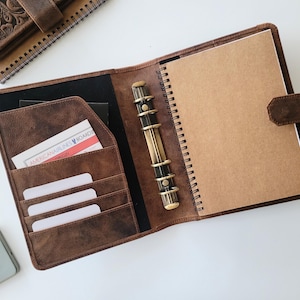 Puede incluir: Carpeta de cuero marr&oacute;n abierta con un cuaderno de espiral y ranuras para tarjetas. La carpeta tiene un mecanismo de anillas color lat&oacute;n y un cierre de correa de cuero. Tambi&eacute;n se ven un bol&iacute;grafo, un llavero de coche y un tel&eacute;fono.