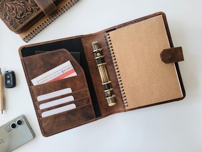 Puede incluir: Carpeta de cuero marr&oacute;n abierta con un cuaderno de espiral y ranuras para tarjetas. La carpeta tiene un mecanismo de anillas color lat&oacute;n y un cierre de correa de cuero. Tambi&eacute;n se ven un bol&iacute;grafo, un llavero de coche y un tel&eacute;fono.