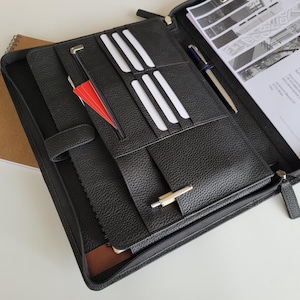 May include: Black leather zipped portfolio with card slots, a pen, and documents. A spiral-bound notebook is in the background. The portfolio is open, showing a red card and white cards.