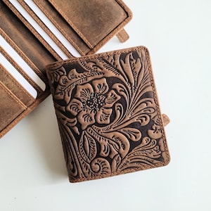 May include: A brown leather wallet with a floral embossed design. The wallet is open, revealing several card slots with white cards. A second wallet is partially visible, also brown leather. The wallets are on a white surface.
