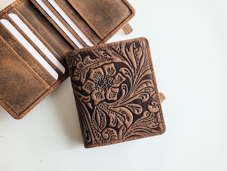 May include: A brown leather wallet with a floral embossed design. The wallet is open, revealing several card slots with white cards. A second wallet is partially visible, also brown leather. The wallets are on a white surface.