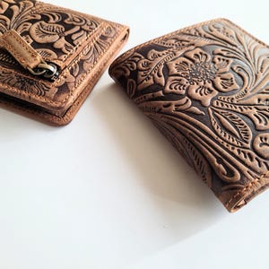 May include: Two brown leather wallets with floral embossed designs. One wallet is closed and has a zipper, while the other is open, showcasing the intricate embossed floral pattern. The wallets are on a white surface.