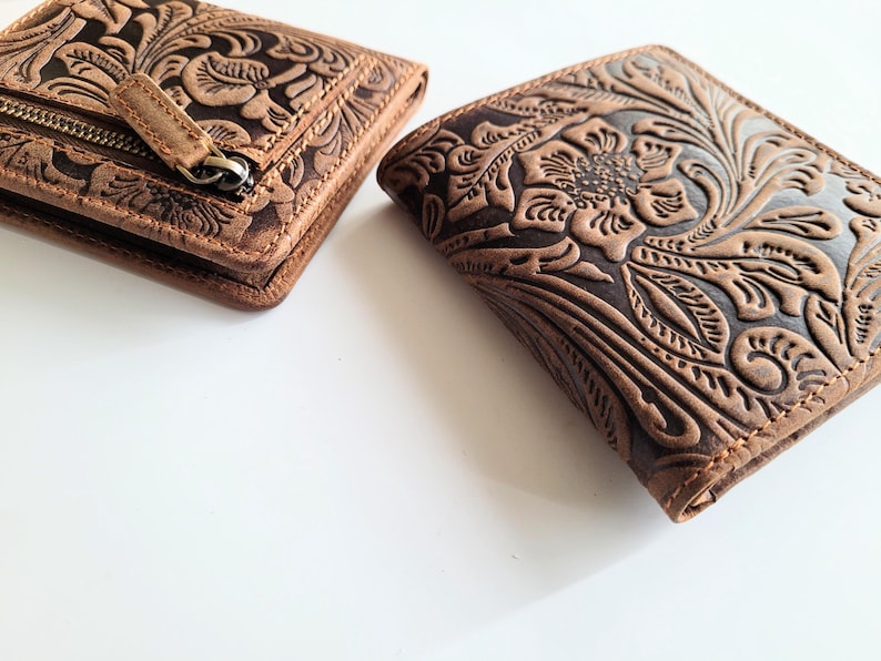 May include: Two brown leather wallets with floral embossed designs. One wallet is closed and has a zipper, while the other is open, showcasing the intricate embossed floral pattern. The wallets are on a white surface.