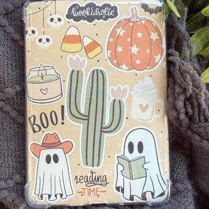 May include: A clear protective case decorated with Halloween-themed stickers. The stickers include a pumpkin, ghosts, a cactus, candy corn, and the text "bookaholic", "BOO!", and "reading time".
