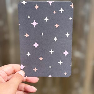 May include: A rectangular card with rounded corners, held by a hand. The card has a dark gray background with a pattern of white, pink, and peach-colored star shapes. The stars vary in size and are evenly distributed across the surface.