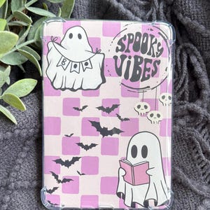 May include: A clear case featuring a pink and white checkered pattern with Halloween-themed illustrations. The design includes ghost figures, bats, and the phrase "Spooky Vibes". The case is designed to fit a tablet or e-reader.