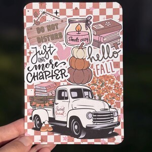 May include: A decorative fall-themed sticker with a white truck carrying books, pumpkins, and the words "book haul." Other text includes "Do Not Disturb," "Just one more chapter," and "Hello Fall." The background is a checkered pattern.