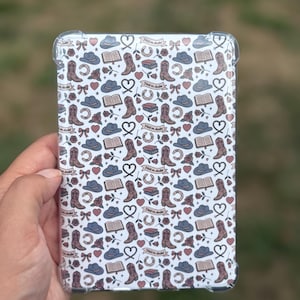 May include: A clear tablet case with a repeating pattern of cowboy boots, hats, hearts, horseshoes, roses, and books. The design includes the text "Hold on darlin'" on a banner. The case is held in a hand.