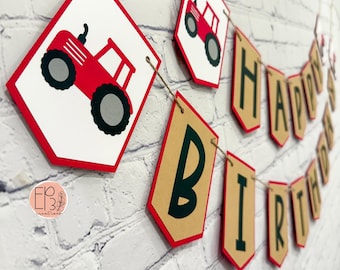 May include: A banner with red and white diamond-shaped pendants featuring a red tractor design. The banner spells out "HAPPY BIRTHDAY" with brown and green letters, strung on twine, against a white brick wall.