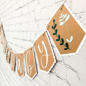 Op de afbeelding: Een bruine papieren banner met witte gegolfde randen en groene en lichtgroene bloemmotieven. De banner heeft de tekst "Oh Baby" in witte cursieve letters.