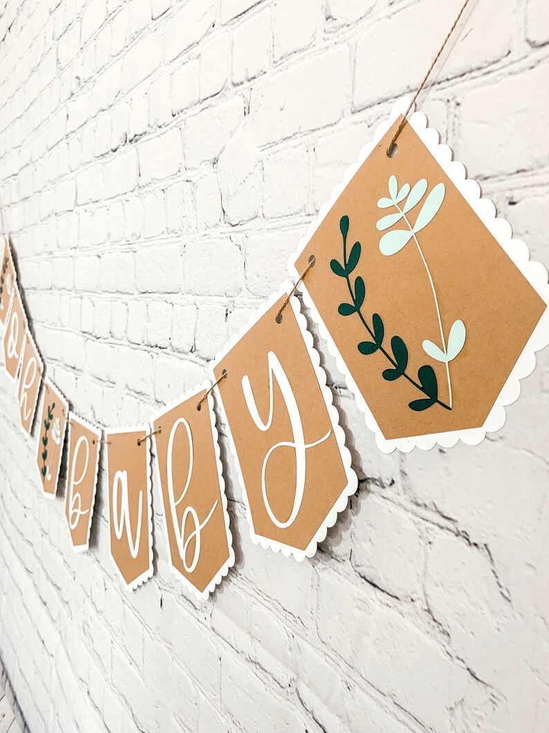 Op de afbeelding: Een bruine papieren banner met witte gegolfde randen en groene en lichtgroene bloemmotieven. De banner heeft de tekst "Oh Baby" in witte cursieve letters.
