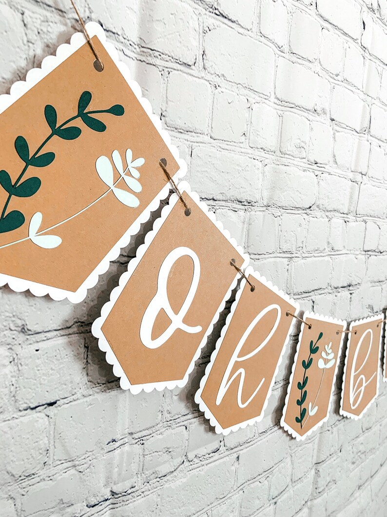 Op de afbeelding: Een bruine papieren banner met witte gegolfde randen en groene en witte bloemenontwerpen. De banner heeft de letters "Oh Baby" in wit erop.