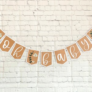 Op de afbeelding: Een bruine papieren banner met witte gegolfde randen en groene bloemmotieven met de tekst "Oh Baby".