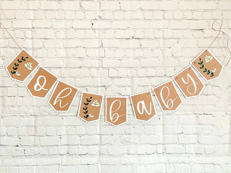 Op de afbeelding: Een bruine papieren banner met witte gegolfde randen en groene bloemmotieven met de tekst "Oh Baby".
