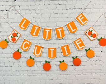 May include: Orange and white banner with the words "Little Cutie" strung on a twine with orange circle cutouts with green leaves.