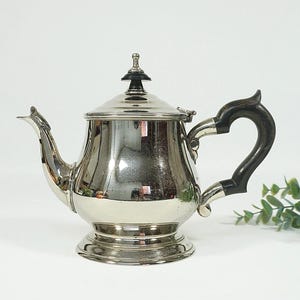 Op de afbeelding: Een zilverkleurige theepot met een zwarte handgreep en dekselknop. De theepot heeft een afgeronde body, een gebogen tuit en een decoratieve voet. De handgreep is van donker hout. De theepot staat op een witte ondergrond.