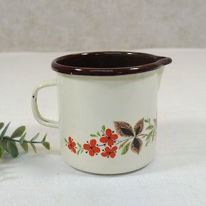 Op de afbeelding: Crèmekleurige emaille kan met een bruine rand en handvat. De kan heeft een bloemenontwerp met rode bloemen en bruine bladeren. De kan heeft een tuit om te schenken.