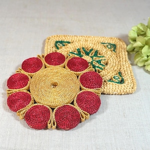 Può includere: Un set di due sottobicchieri intrecciati. Uno è rotondo con una spirale centrale e dettagli rossi, l'altro è quadrato con ricami floreali verdi. Entrambi sono realizzati con fibre naturali e adatti all'arredamento della casa.