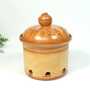 Pode incluir: Um recipiente de cerâmica para cebolas com tampa. O recipiente é bege claro com uma tampa e um botão marrom escuro. A palavra "ZWIEBEL" está em relevo na tampa. Possui orifícios para ventilação.